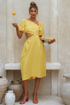 Woman wearing a yellow linen midi dress with puffed sleeves and asymmetrical cut-out design standing in a room with decorative vases, tossing a lemon and holding a plate of lemons.