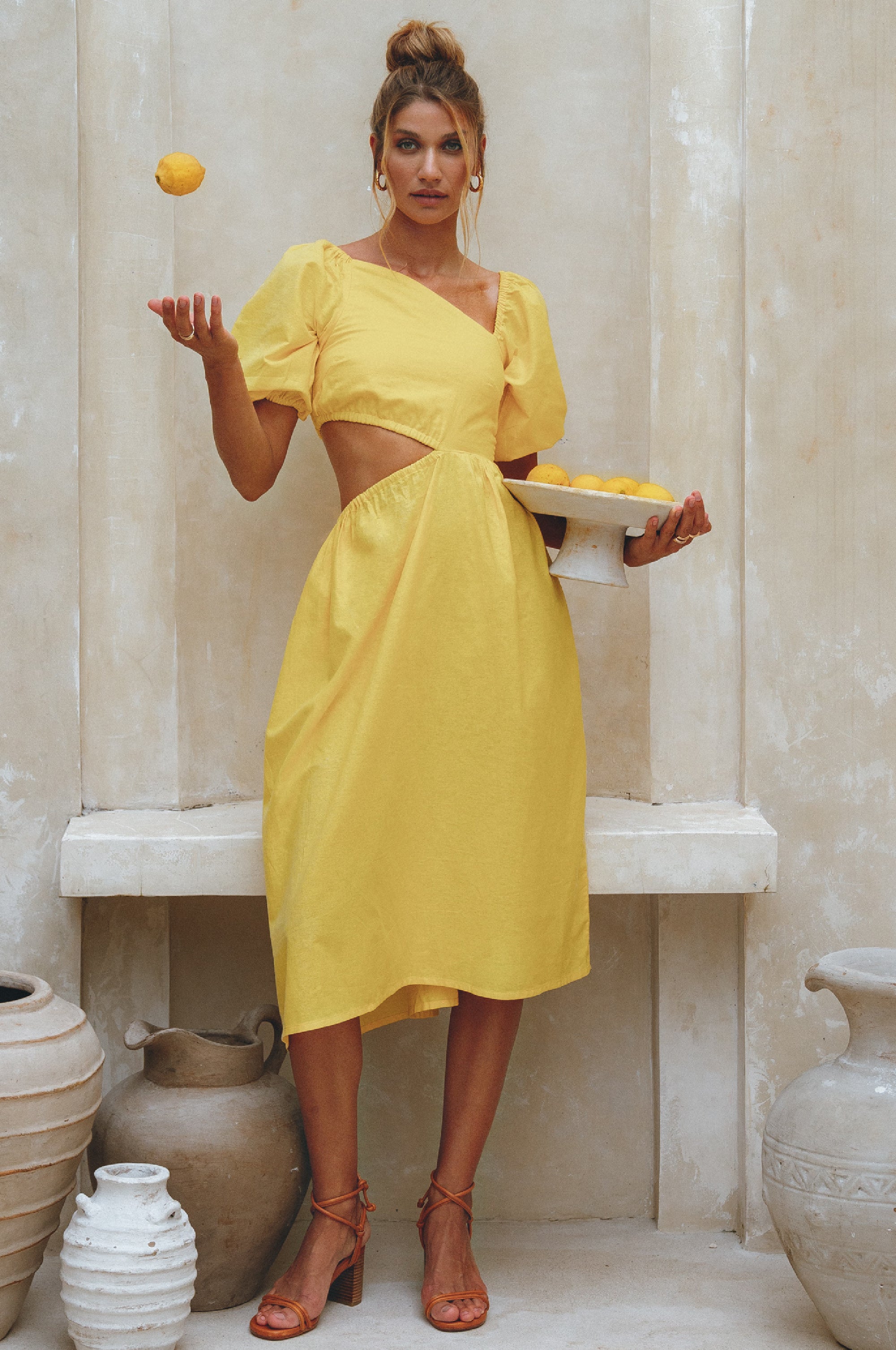 Woman wearing a yellow linen midi dress with puffed sleeves and asymmetrical cut-out design standing in a room with decorative vases, tossing a lemon and holding a plate of lemons.