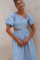 Woman wearing a light blue linen midi dress with puffed sleeves and asymmetrical cut-out design against a beige background