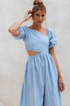 Woman wearing a light blue linen midi dress with puffed sleeves and asymmetrical cut-out design against a plain background