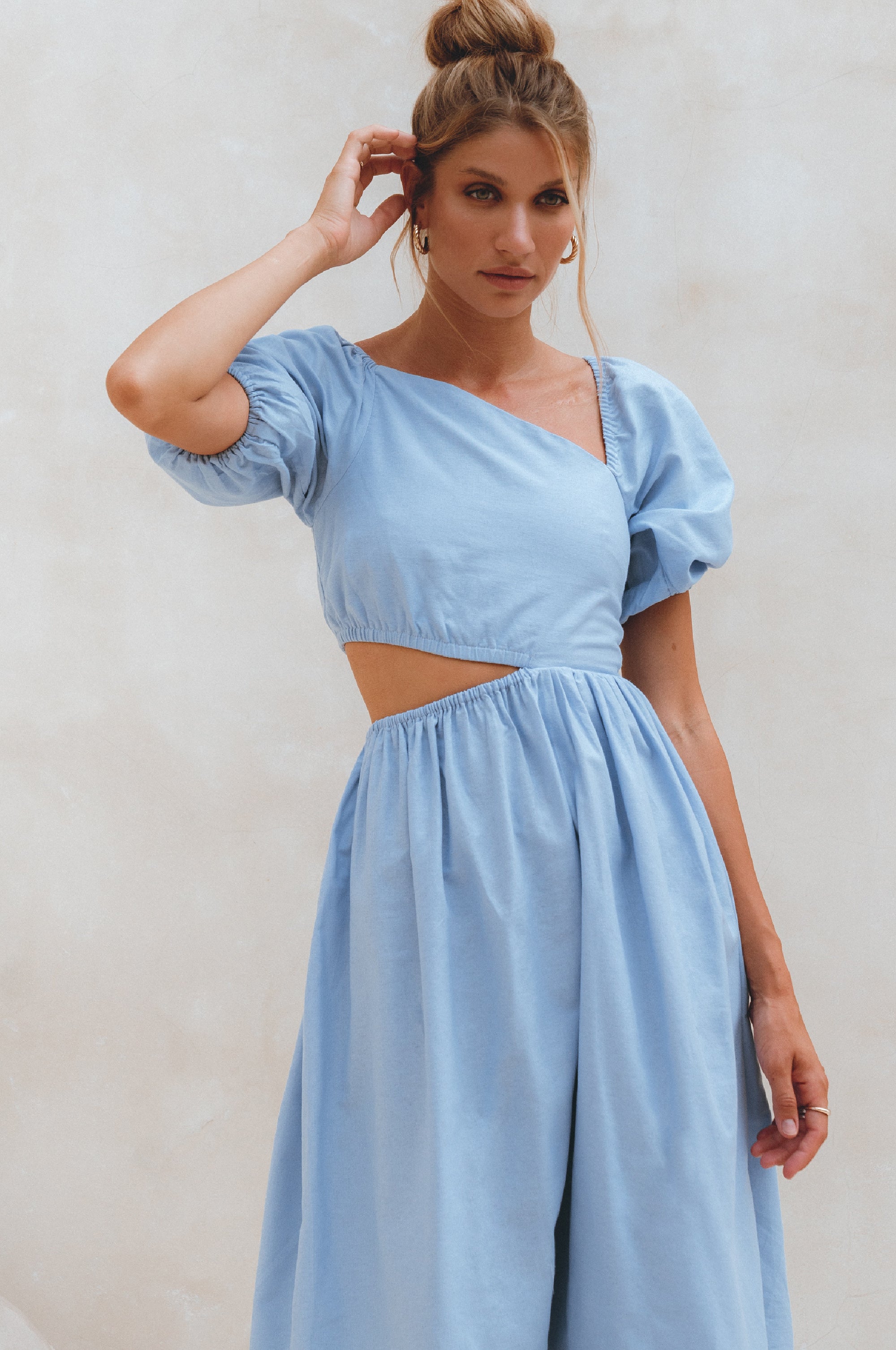 Woman wearing a light blue linen midi dress with puffed sleeves and asymmetrical cut-out design against a plain background