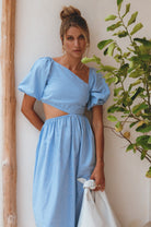 Woman wearing a light blue linen midi dress with puffed sleeves and asymmetrical cut-out design, standing next to a plant.