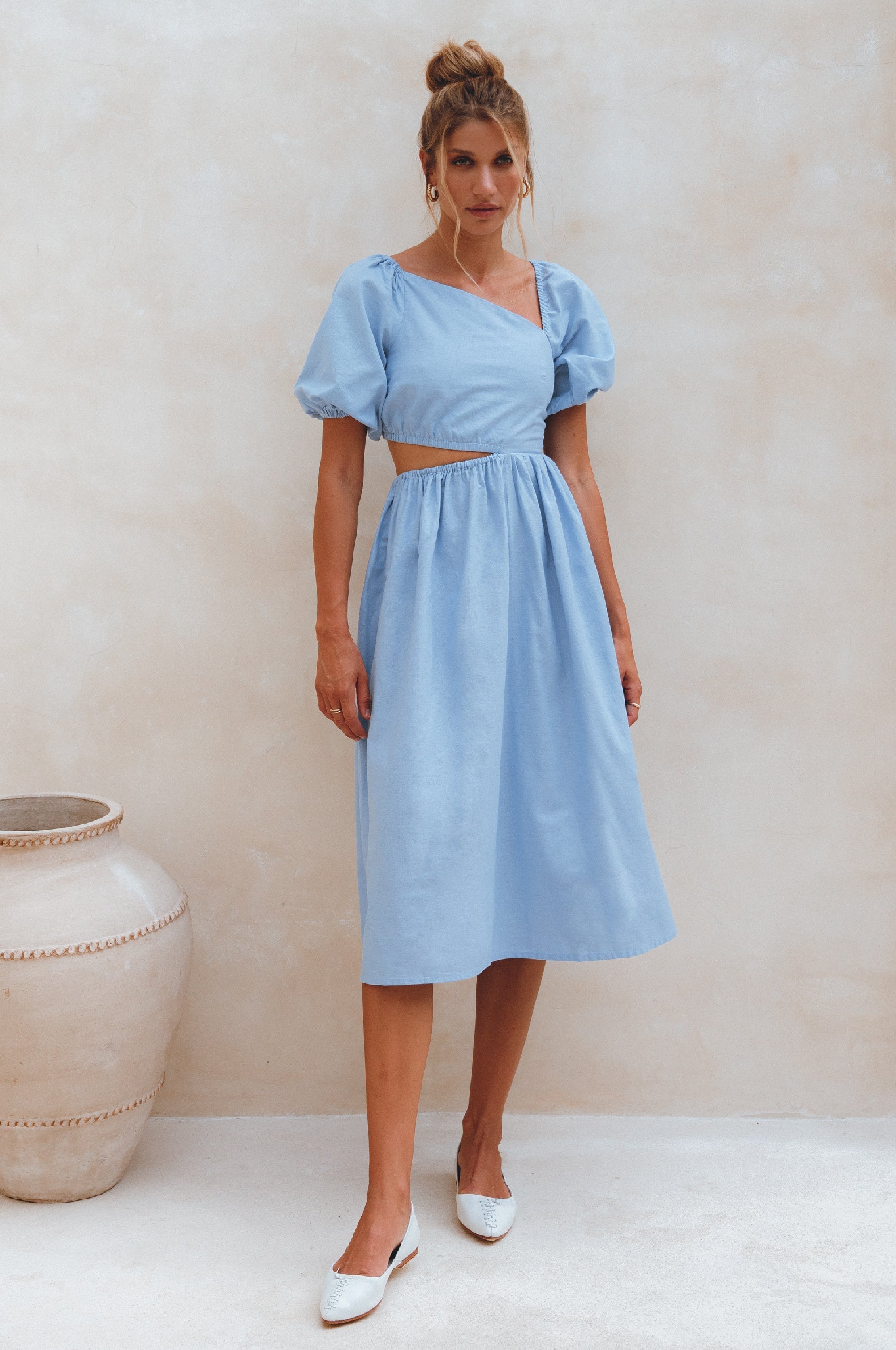 Woman wearing a light blue linen midi dress with puffed sleeves and asymmetrical cut-out design standing against a plain background, front view.