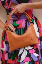 Person holding a tan leather handbag with a colorful dress in the background