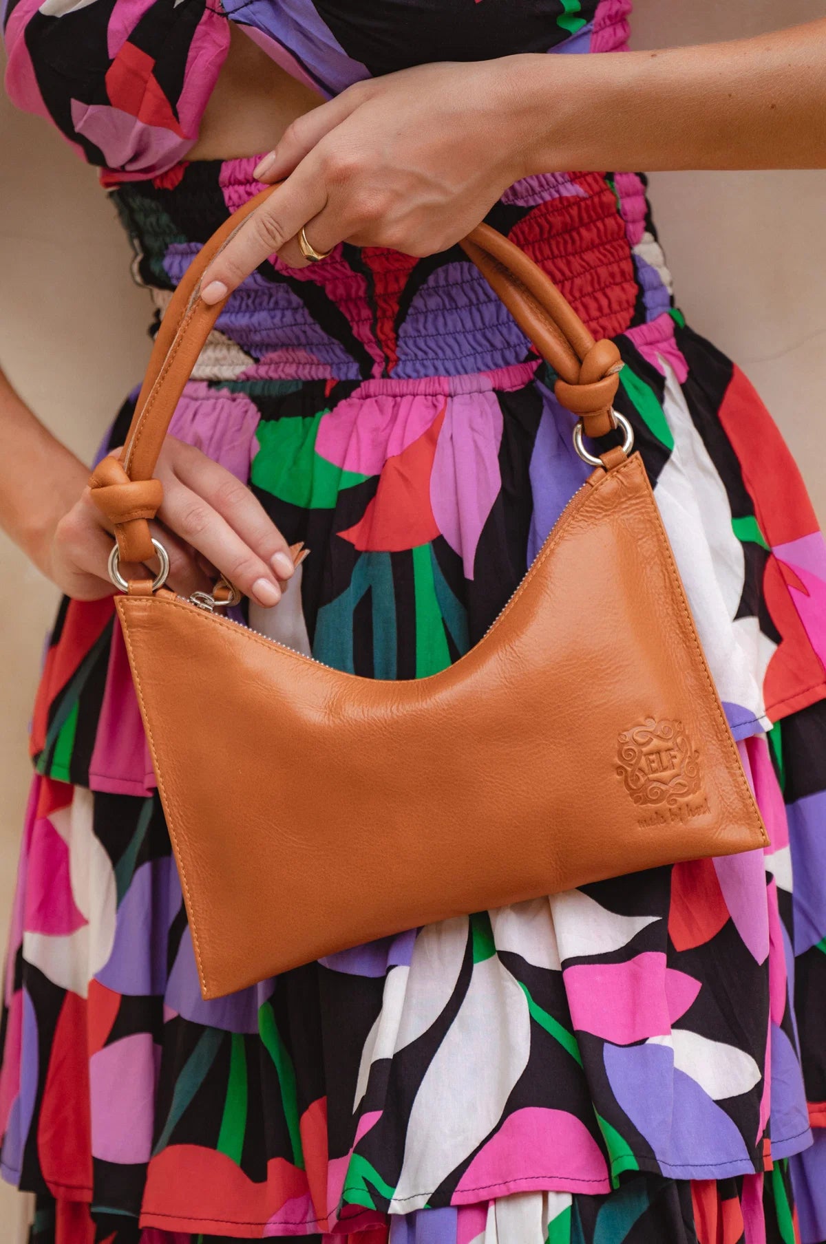 Person holding a tan leather handbag with gold hardware and knotted leather handle with a colorful dress in the background
