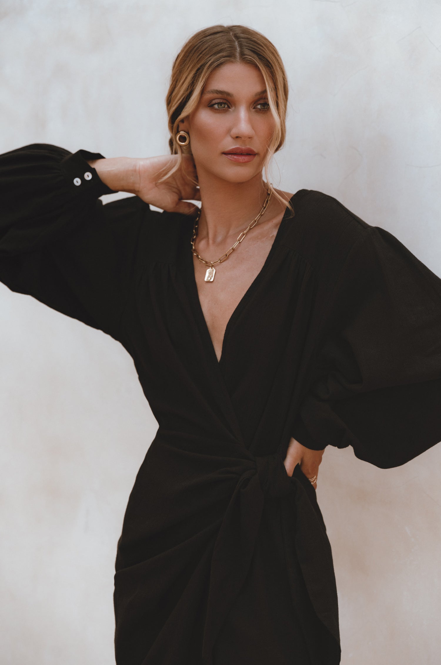 Woman wearing a black crinkle linen mini dress with adjustable wrap, a V-neckline and billowy sleeves, against a white background, front view close up