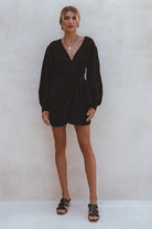 Woman wearing a black crinkle linen mini dress with adjustable wrap, a V-neckline and billowy sleeves, against a white background, front view