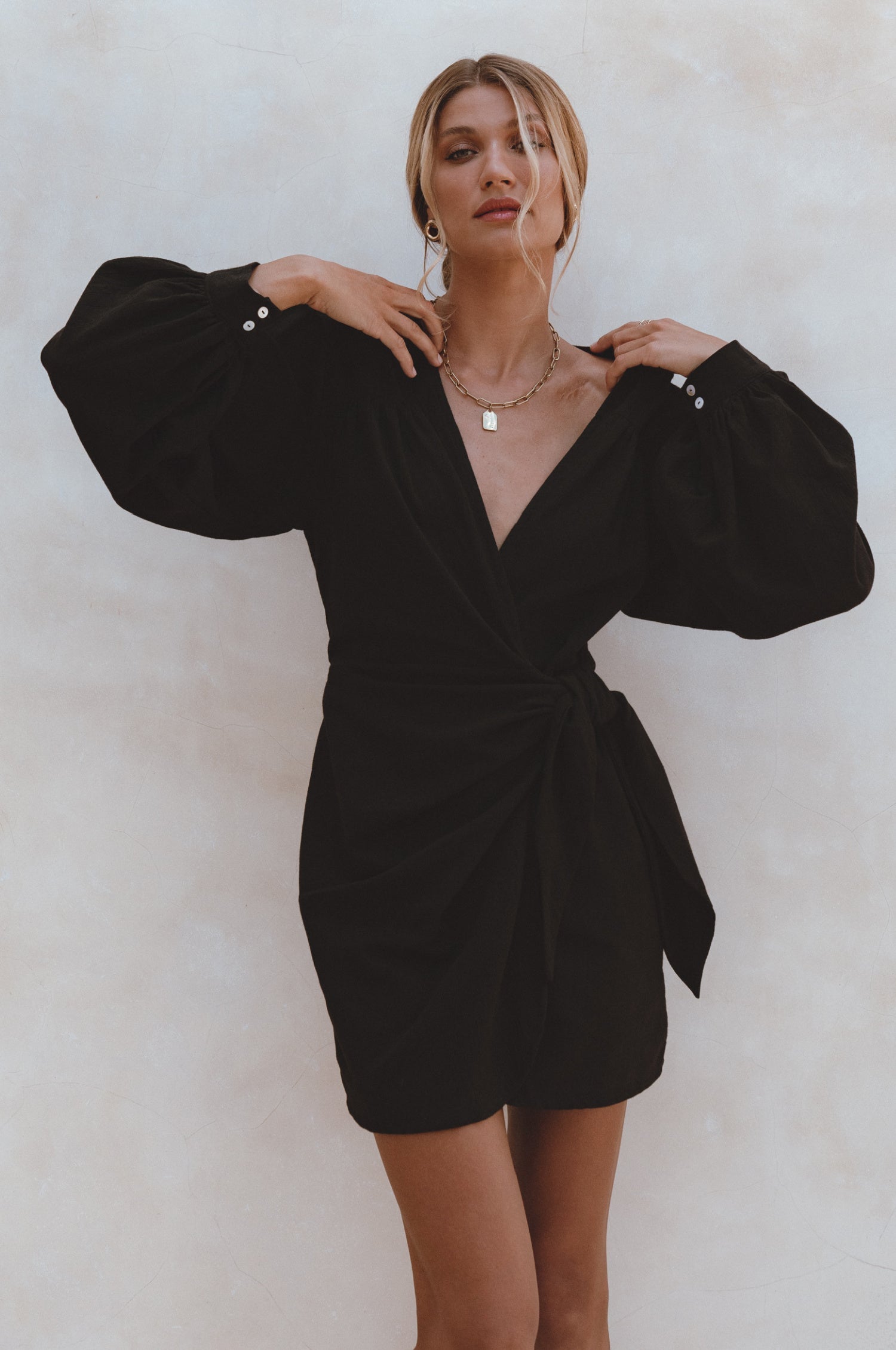 Woman wearing a black crinkle linen mini dress with adjustable wrap, a V-neckline and billowy sleeves, against a white background, front view
