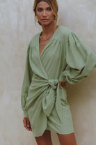 Woman wearing a sherbert green crinkle linen mini dress with adjustable wrap, a V-neckline and billowy sleeves, against a neutral background, front view 