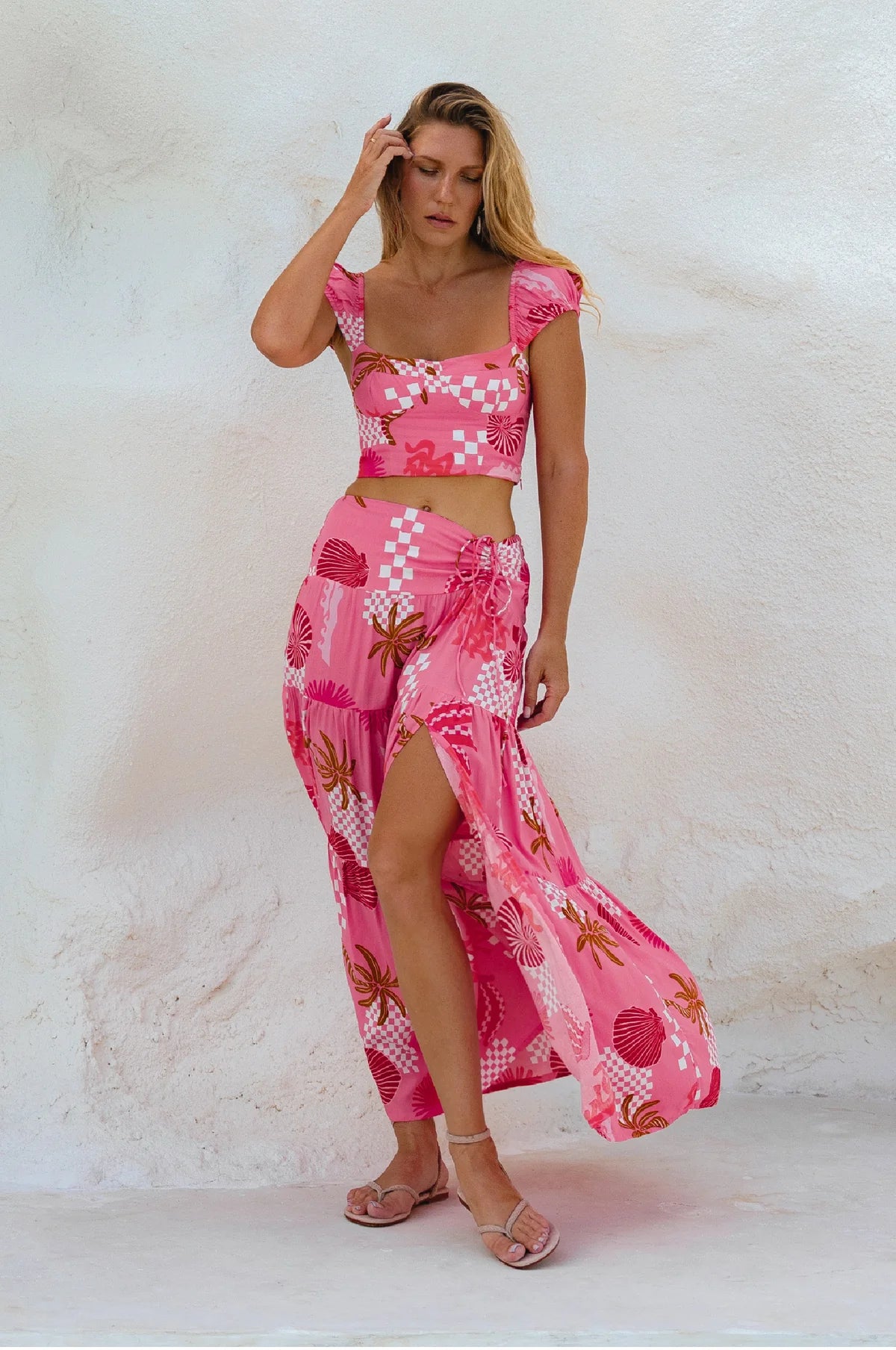 A woman modeling a pink two-piece tropical print set consisting of a crop top and a matching high-slit maxi skirt, standing against a textured white stucco wall.