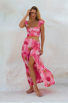 A woman modeling a pink two-piece tropical print set consisting of a crop top and a matching high-slit maxi skirt, standing against a textured white stucco wall.