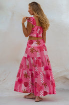 A woman wearing a bright pink bohemian-style two-piece set with a crop top and a tiered maxi skirt, photographed against a textured white wall, back view