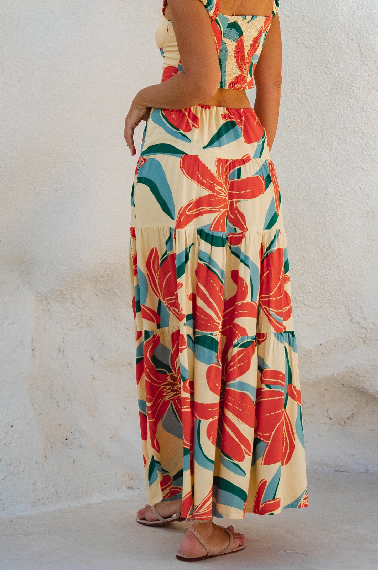 Woman wearing a floral two-piece cropped top with maxi skirt with red and green patterns on a white background, back view
