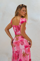 Woman wearing a pink, white and magenta abstract floral and shell motif cropped top with flutter sleeve and matching skirt with white background, back view