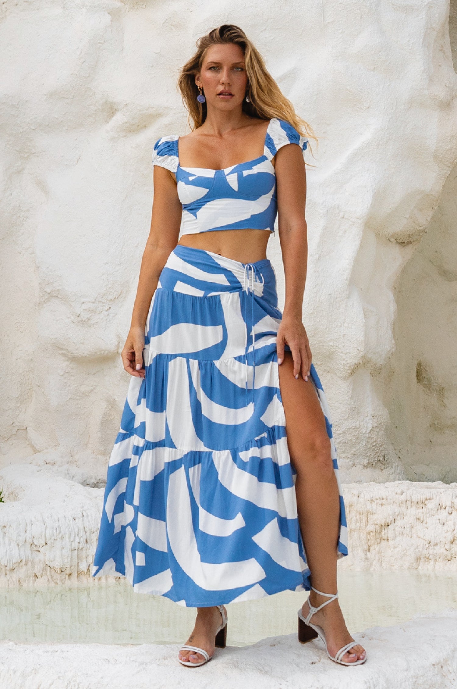 Woman wearing a blue and white patterned matching two-piece cropped top and maxi skirt with high slit against a textured white background
