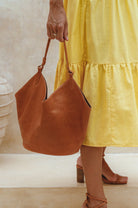 Person holding a tan soft leather handbag wearing a yellow dress and brown shoes.