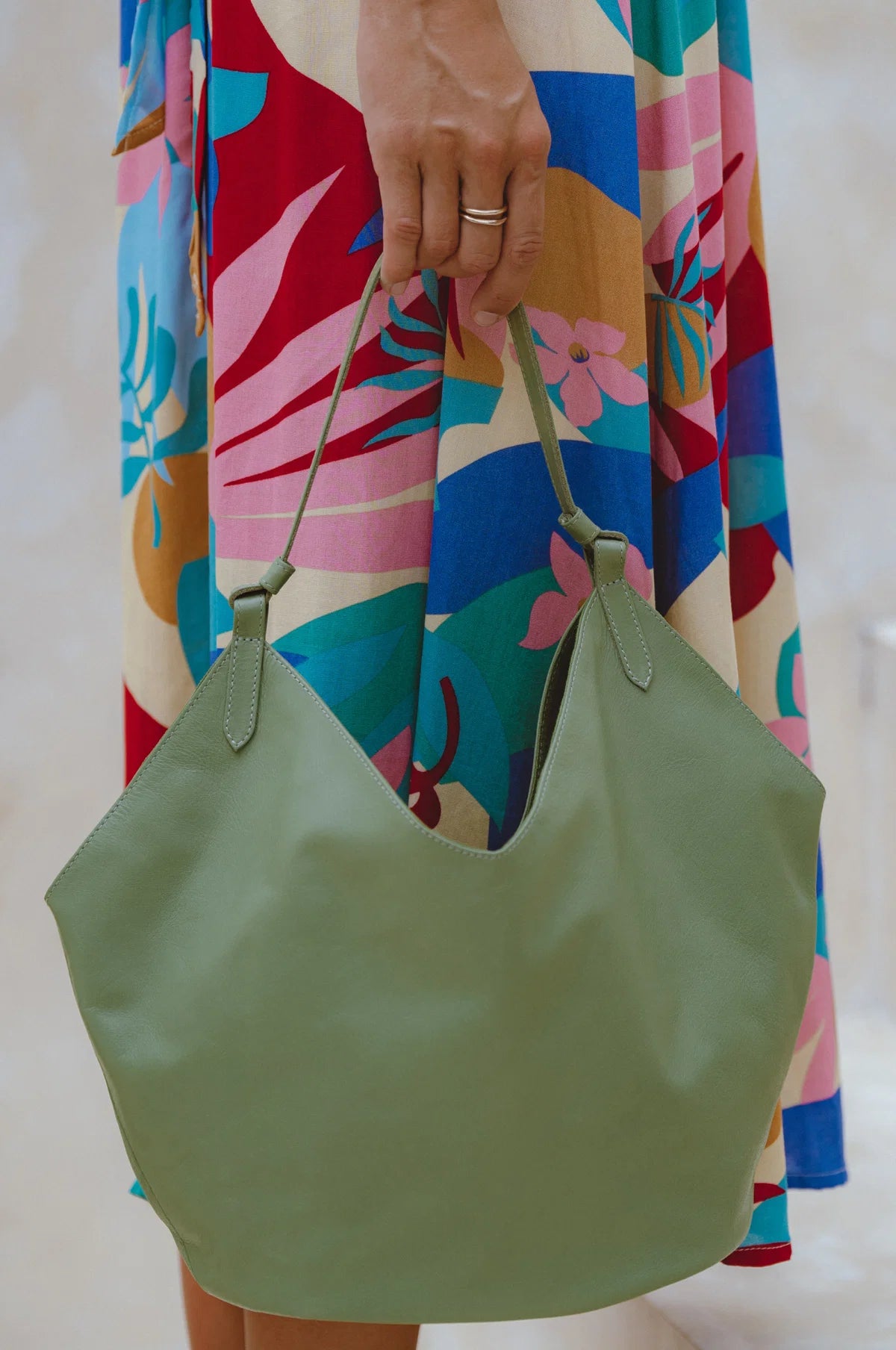 A person holding a large green leather hobo tote bag, wearing a colorful tropical print dress. The bag has a single shoulder strap and is shown against a light, neutral background.