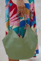 A person holding a large green leather hobo tote bag, wearing a colorful tropical print dress. The bag has a single shoulder strap and is shown against a light, neutral background.