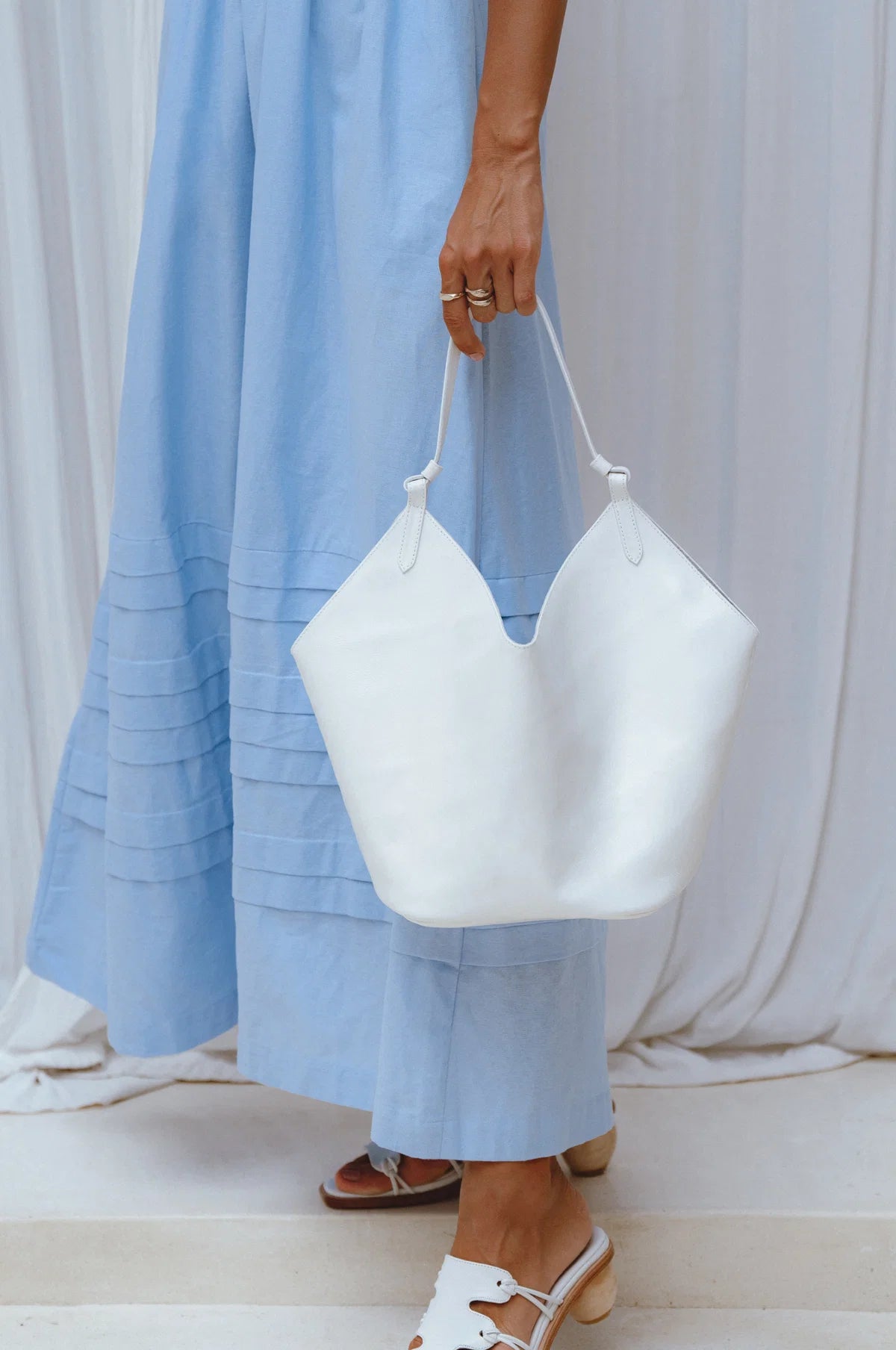 A person wearing a light blue dress holds a large white leather handbag with single leather handle. The person is also wearing white sandals, standing in front of a draped white fabric background.