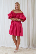 Woman wearing a bright pink off-shoulder dress against a white curtain background