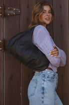 Woman wearing a large black leather slouchy bag and light purple sweater against a wooden door.