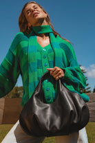 A woman outdoors wearing a green striped knit cardigan and holding a large black leather slouchy handbag, with a blue sky and wooden fence in the background.