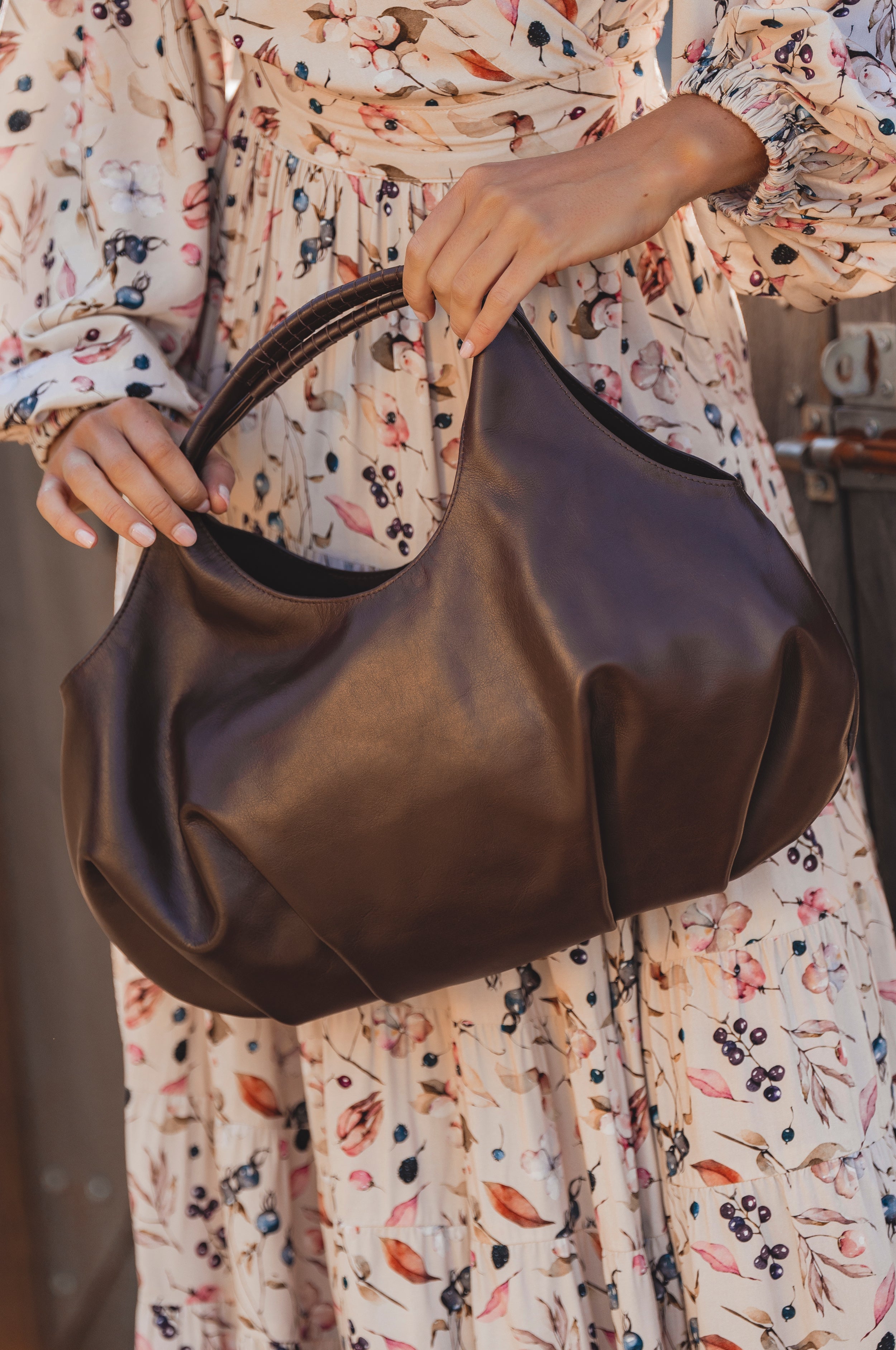 Person holding a dark chocolate brown leather handbag wearing a floral dress.