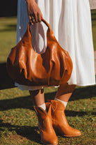 Person wearing a tan leather handbag and boots standing on grass