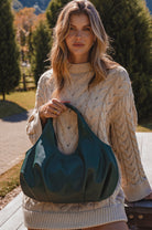 Woman holding a green leather bag outdoors with trees and a path in the background
