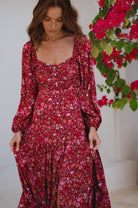 Woman wearing a red floral dress standing next to a plant with red flowers.
