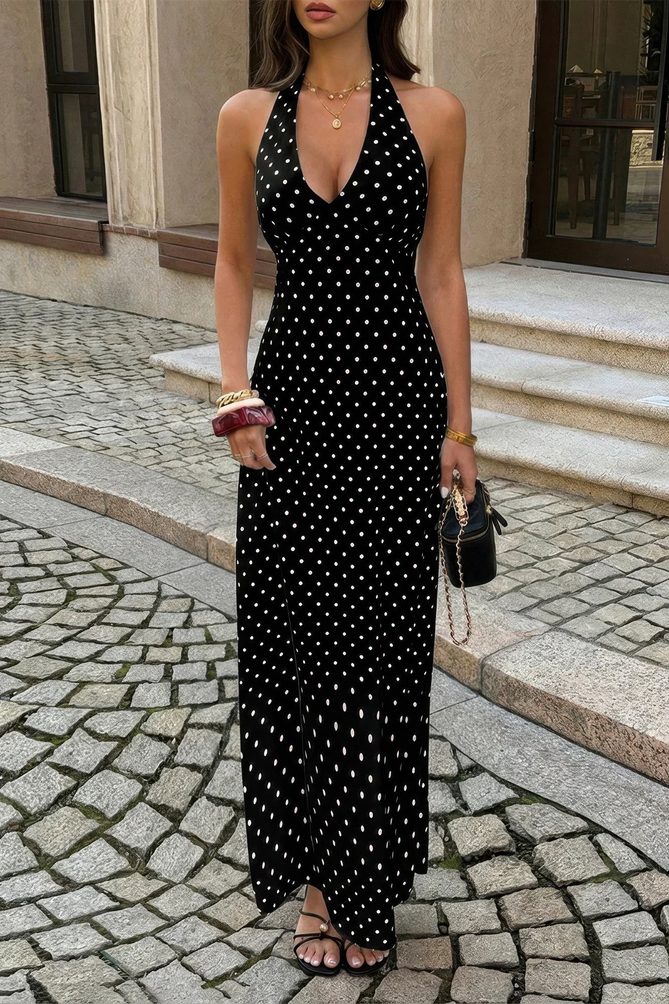 Woman wearing black long with maxi dress with white polka dots with halter and deep v-neckline, standing in a cobbled walkway  in front of a store window
