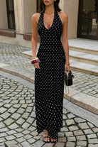 Woman wearing black long with maxi dress with white polka dots with halter and deep v-neckline, standing in a cobbled walkway  in front of a store window