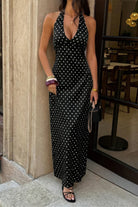 Woman wearing long black with maxi dress with white polka dots with halter and deep v-neckline, standing in front of restaurant window.