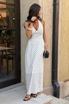 Woman wearing long white with maxi dress with black polka dots with halter and deep v-neckline, against a store window