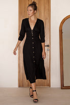 Woman wearing a black v-neck midi dress with buttons standing in front of a wooden door.