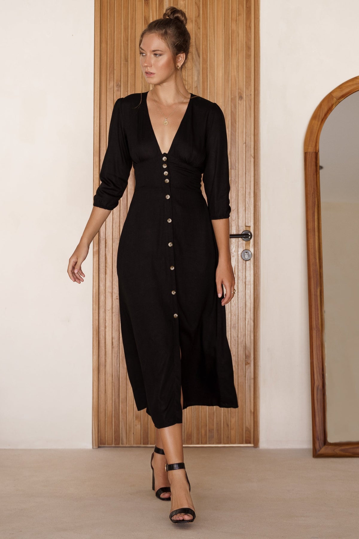 Woman wearing a black v-neck midi dress with buttons standing in front of a wooden door.