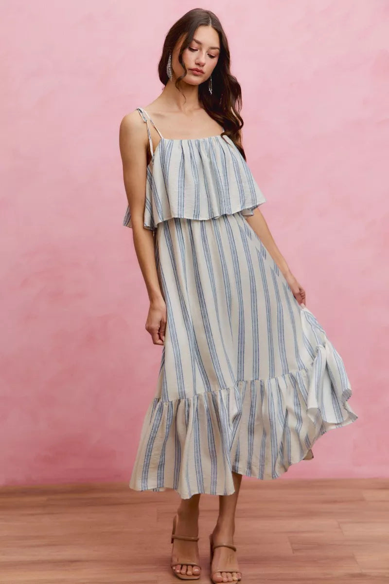 Woman wearing a blue stripe maxi dress with adjustable tie shoulders, a flowing overlay bodice, and a ruffle hem.
