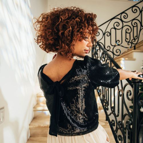 Woman wearing a black sequin top with a back bow tie, photographed on a staircase for the holiday collection.