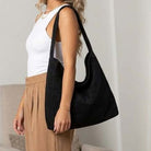 A woman wearing a white tank top and beige pleated pants carries a large black suede hobo shoulder bag in a bright indoor setting.