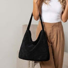 A woman wearing a white tank top and tan pants carries a large black suede hobo shoulder bag.