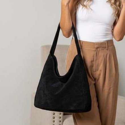 A woman wearing a white tank top and tan pants carries a large black suede hobo shoulder bag.