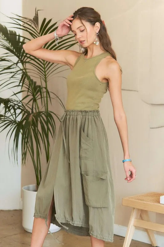 Model wearing an olive green mixed media cargo midi dress with a ribbed tank bodice, cargo pockets, drawstring waist, and adjustable drawstring hem.
