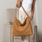 A woman wearing a white tank top and beige pants carries a large tan suede hobo shoulder bag in an indoor setting with a beige armchair.