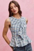 Woman wearing a blue plaid sleeveless peplum top with ruched detailing and a cascading front ruffle.
