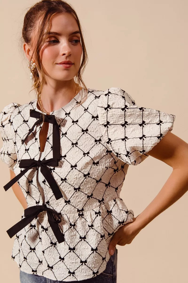 Close up of textured cream bow front top featuring black ribbon ties, puff sleeves, and peplum silhouette | AVAH Collections