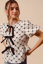Close up of textured cream bow front top featuring black ribbon ties, puff sleeves, and peplum silhouette | AVAH Collections