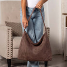 A person wearing light blue jeans holds a large brown suede hobo shoulder bag in a living room setting with a beige armchair in the background.